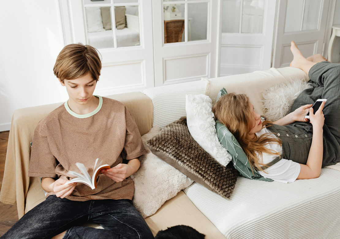 To teens, en dreng og pige i en sofa. Dreng sidder og læser en bog, pige ligger med telefon