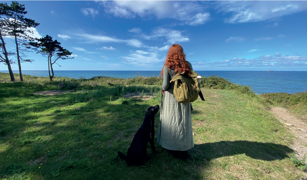Freja Oschlak står med  rygsæk på og ryggen til og kigger ud over vandet. Ved hendes side sidder en sort hund.