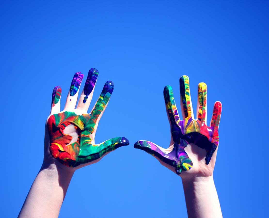 Hænder løftet i vejret, så håndfladerne vender mod beskueren. Håndfladerne er dækket af adskillige forskellige farver maling.
