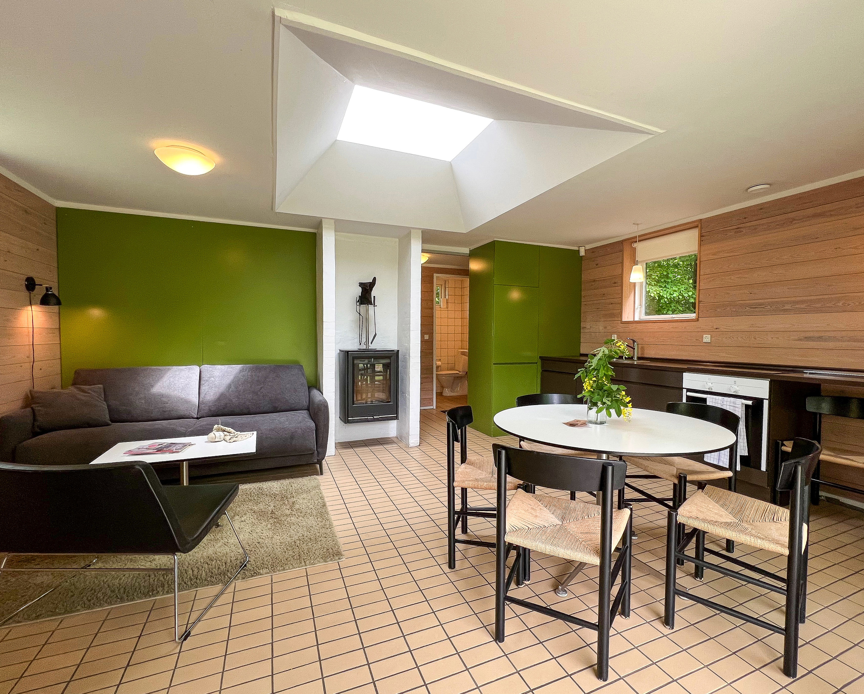 Køkken -alrum med køkken i rummets højre side. Sofa, spisebord og pejs. Grøn bagvæg og ovenlysvindue. Flisegulv.