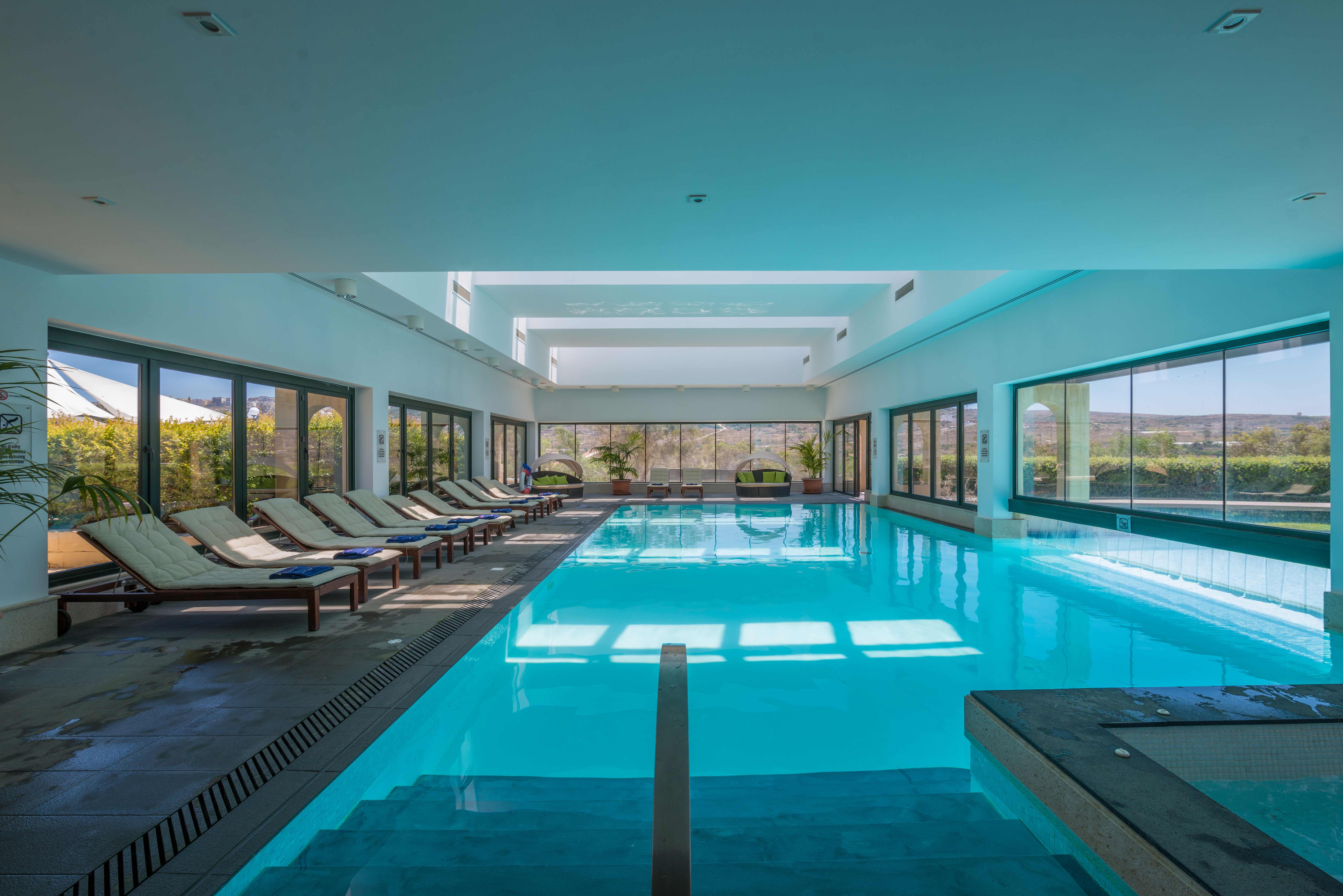 Indendørs poolområde på Mellieha Holiday Centre. Pool med liggestole rundt langs store vinduer.