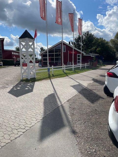 Indgang til campingplads. Rød bygning i baggrund, delvis parkeringsplads og bom i forgrund